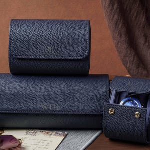 May include: Three navy blue textured watch rolls with a pebbled finish. Two rolls are closed, and one is open, revealing a watch inside. The rolls feature gold-tone snap buttons and embossed lettering on the front flaps, including WDL.