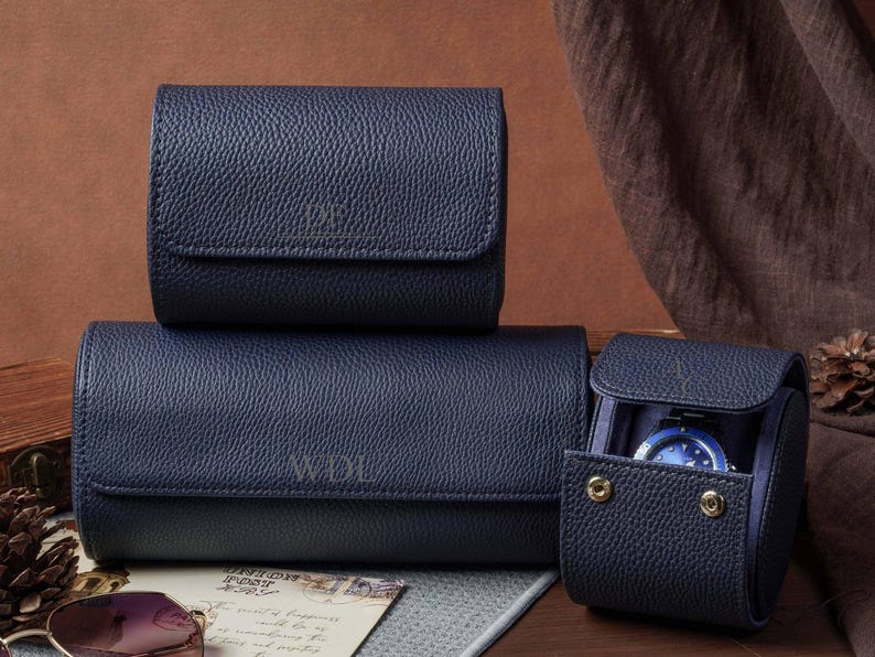 May include: Three navy blue textured watch rolls with a pebbled finish. Two rolls are closed, and one is open, revealing a watch inside. The rolls feature gold-tone snap buttons and embossed lettering on the front flaps, including WDL.