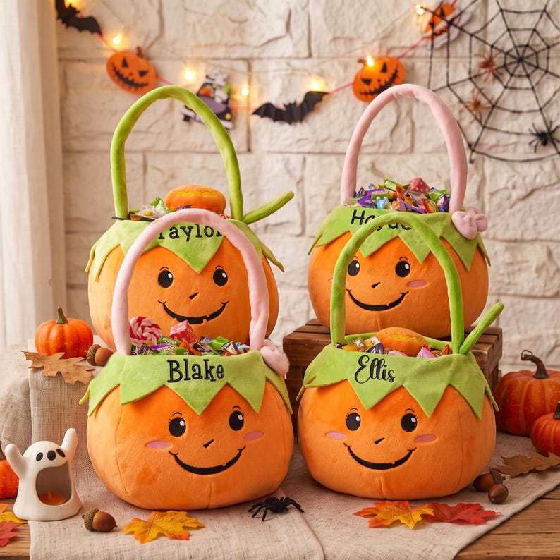 May include: Four plush pumpkin-shaped Halloween candy buckets with green leaf-shaped rims and handles. Each bucket features a smiling face and is embroidered with a name: Taylor, Hayden, Blake, or Ellis. The buckets are filled with assorted candies.