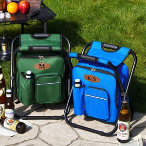 May include: Two portable cooler chairs, one green and one blue, each featuring a black metal frame, a zippered main compartment, a front pocket with a reflective strip, a side bottle holder, and a brown leather-like patch with initials.
