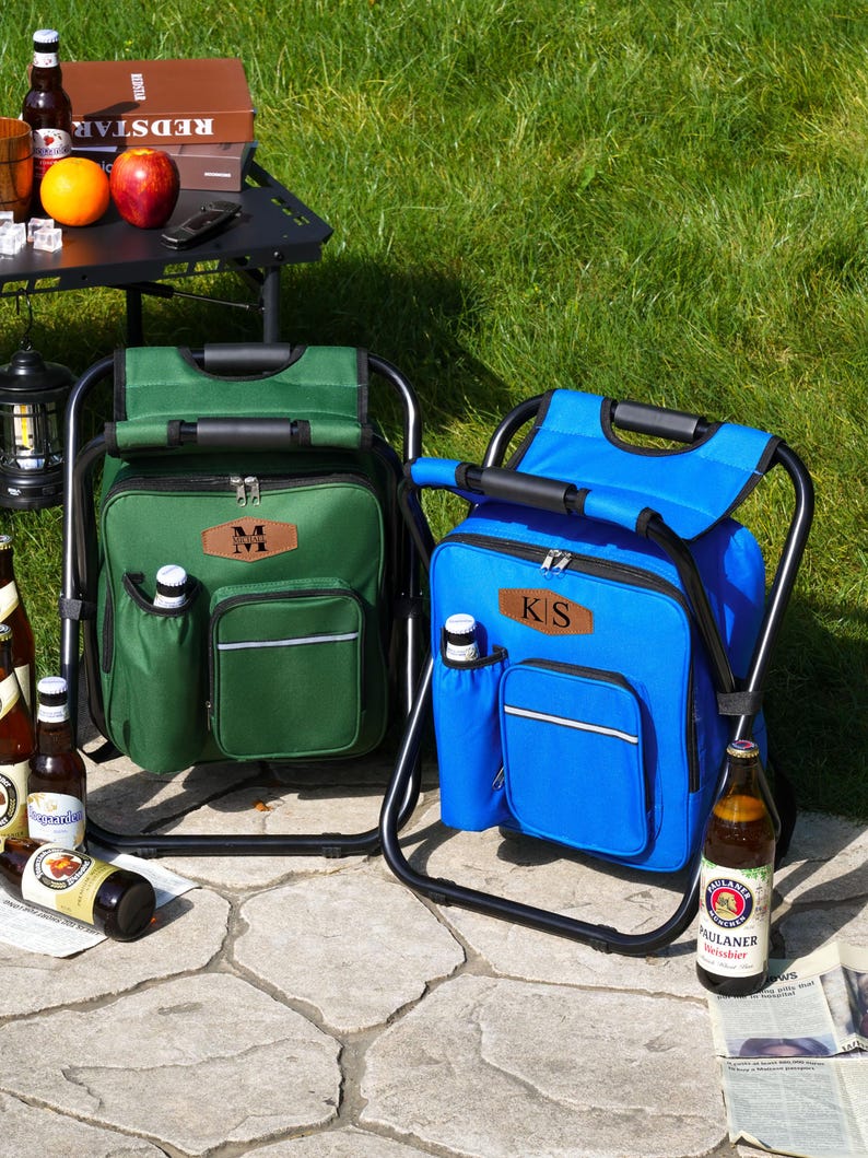 May include: Two portable cooler chairs, one green and one blue, each featuring a black metal frame, a zippered main compartment, a front pocket with a reflective strip, a side bottle holder, and a brown leather-like patch with initials.