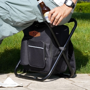 May include: A black folding stool with an integrated fabric storage bag featuring a brown leather-like patch with the letter S. The stool has a sturdy black metal frame and a front pocket with a horizontal reflective silver strip.