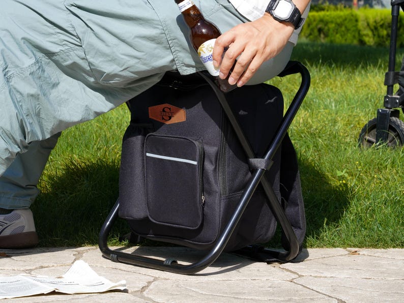 May include: A black folding stool with an integrated fabric storage bag featuring a brown leather-like patch with the letter S. The stool has a sturdy black metal frame and a front pocket with a horizontal reflective silver strip.