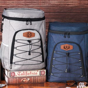 May include: Two insulated cooler backpacks, one gray and one navy blue, with black accents and front bungee cords. The gray backpack has a leather patch with the letter "D", and the blue one has the letter "R".