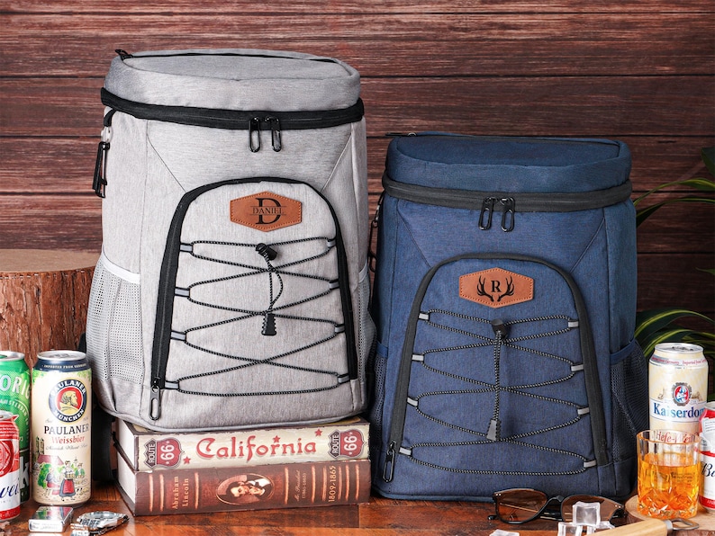 May include: Two insulated cooler backpacks, one gray and one navy blue, with black accents and front bungee cords. The gray backpack has a leather patch with the letter "D", and the blue one has the letter "R".