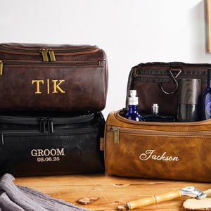 May include: Three personalised leather wash bags in brown and black. One bag is open, displaying blue bottles and grooming products. The bags are embroidered with initials, names, and a date. A wooden shaving brush and razor are on the wooden surface.