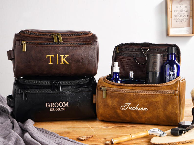May include: Three personalised leather wash bags in brown and black. One bag is open, displaying blue bottles and grooming products. The bags are embroidered with initials, names, and a date. A wooden shaving brush and razor are on the wooden surface.
