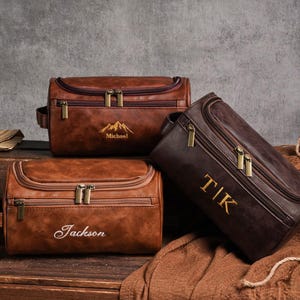 May include: Three brown leather wash bags with gold zips and personalised embroidery. One bag has a mountain design and the name "Michael", another has the name "Jackson", and the third has the initials "TIK".