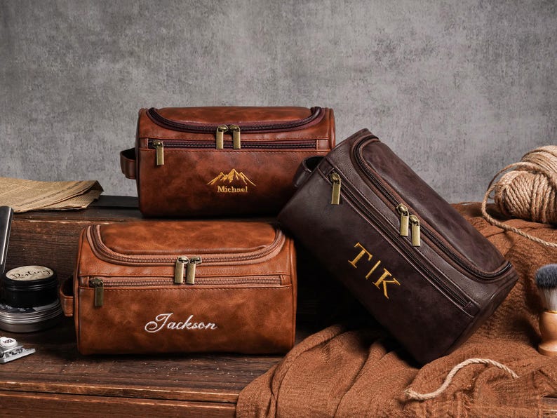 May include: Three brown leather wash bags with gold zips and personalised embroidery. One bag has a mountain design and the name "Michael", another has the name "Jackson", and the third has the initials "TIK".