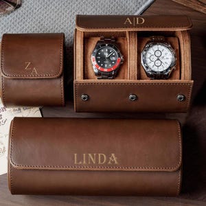 May include: Three brown leather watch cases with snap closures and personalized engravings. One case is closed with the initials ZA, one is open displaying two wristwatches with the initials A|D, and one is closed with the name LINDA engraved.