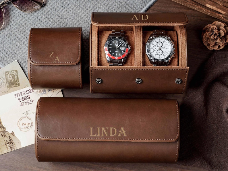 May include: Three brown leather watch cases with snap closures and personalized engravings. One case is closed with the initials ZA, one is open displaying two wristwatches with the initials A|D, and one is closed with the name LINDA engraved.