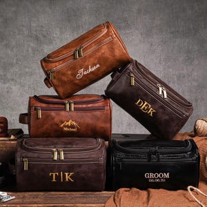 May include: Several personalised leather wash bags in shades of brown and black. Each bag has a zip closure and is decorated with custom text or a mountain graphic. The bags are arranged on a wooden surface.