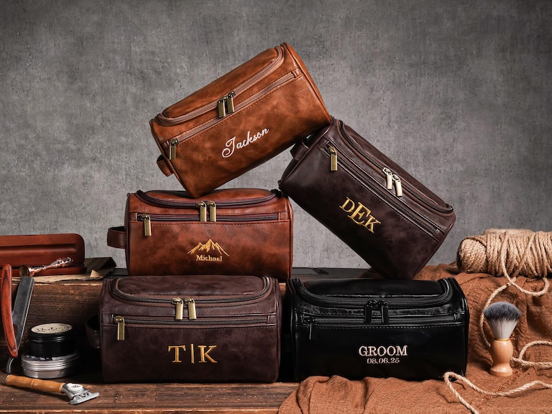 May include: Several personalised leather wash bags in shades of brown and black. Each bag has a zip closure and is decorated with custom text or a mountain graphic. The bags are arranged on a wooden surface.