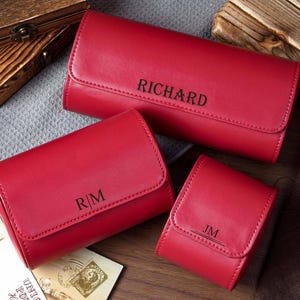 May include: Three red rectangular cases with rounded edges and flap closures. The largest case is engraved with the name RICHARD, the medium case with the initials R|M, and the smallest case with the initials JM, all in a dark, sans-serif font.
