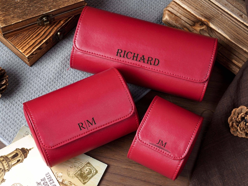 May include: Three red rectangular cases with rounded edges and flap closures. The largest case is engraved with the name RICHARD, the medium case with the initials R|M, and the smallest case with the initials JM, all in a dark, sans-serif font.