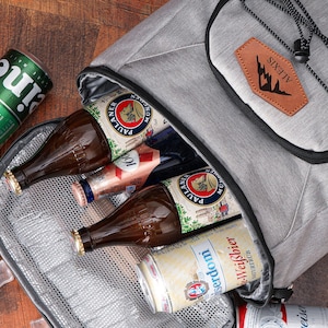 May include: A gray insulated cooler bag with a black zipper, containing beer bottles and cans. The bag has a brown leather patch with the word "ALEXIS" and a mountain silhouette. A green Heineken can and a glass of liquid with ice cubes are visible.