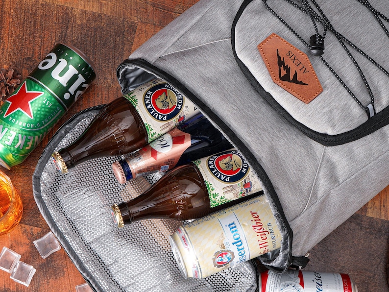 May include: A gray insulated cooler bag with a black zipper, containing beer bottles and cans. The bag has a brown leather patch with the word "ALEXIS" and a mountain silhouette. A green Heineken can and a glass of liquid with ice cubes are visible.