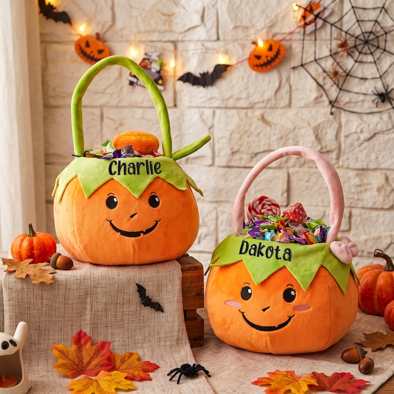 May include: Two plush pumpkin-shaped Halloween candy baskets with green leaf-shaped rims. The left basket features the name Charlie embroidered in black, and the right basket features the name Dakota embroidered in black. Both are filled with candy.