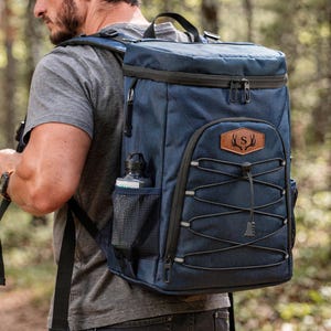 May include: A navy blue backpack with black accents, a brown leather patch with a deer antler design, and a water bottle in a side pocket. The backpack has a drawstring closure and a top handle.