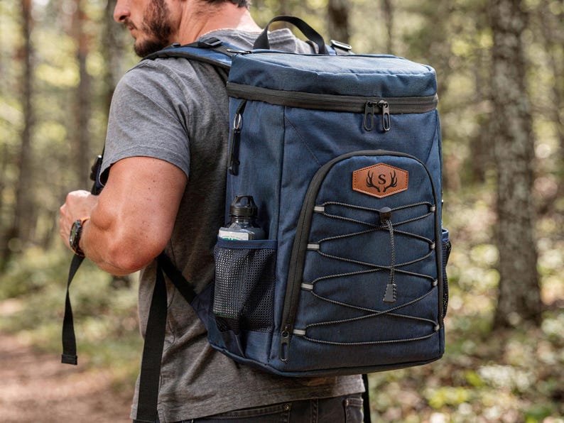May include: A navy blue backpack with black accents, a brown leather patch with a deer antler design, and a water bottle in a side pocket. The backpack has a drawstring closure and a top handle.