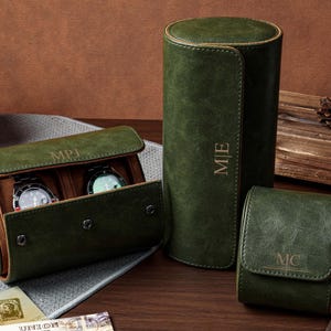 May include: Three dark green cylindrical watch cases with tan interiors. One case is open, revealing two wristwatches. The cases feature gold-embossed monograms reading MPJ, M|E, and MC. The exterior has a textured finish with snap closures.