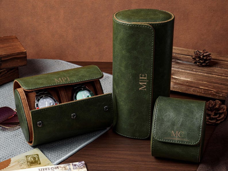 May include: Three dark green cylindrical watch cases with tan interiors. One case is open, revealing two wristwatches. The cases feature gold-embossed monograms reading MPJ, M|E, and MC. The exterior has a textured finish with snap closures.