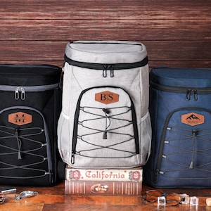 May include: Three insulated cooler backpacks in black, gray, and navy blue. Each has a front pocket with a leather patch displaying initials or a mountain logo. The backpacks have black zippers and a bungee cord design.