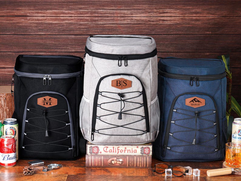 May include: Three insulated cooler backpacks in black, gray, and navy blue. Each has a front pocket with a leather patch displaying initials or a mountain logo. The backpacks have black zippers and a bungee cord design.