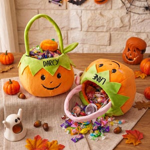 May include: Two plush pumpkin-shaped candy baskets with green leaf-shaped rims. One basket is upright with the name DARCY embroidered in black, and the other is tilted on its side with the name LANE embroidered in black, spilling assorted candies.