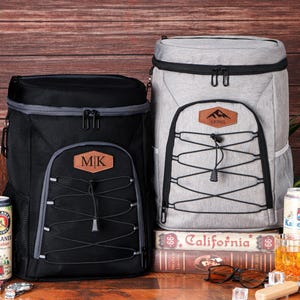 May include: Two insulated cooler backpacks, one black and one gray, with personalized leatherette patches. The black backpack has "MK" on its patch. The gray backpack has a mountain logo and the name "Lionel". Cans of beer and a glass of amber liquid are visible.