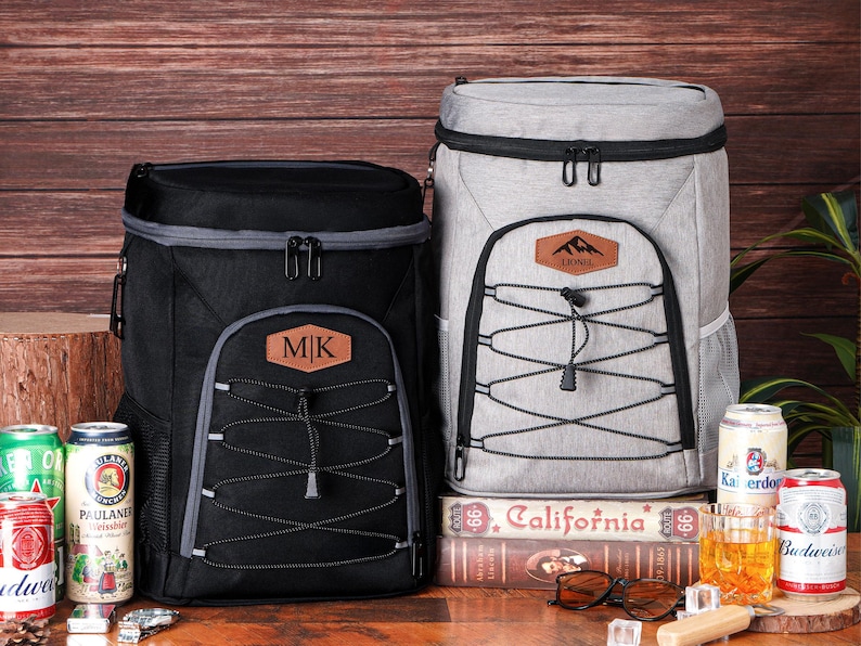 May include: Two insulated cooler backpacks, one black and one gray, with personalized leatherette patches. The black backpack has "MK" on its patch. The gray backpack has a mountain logo and the name "Lionel". Cans of beer and a glass of amber liquid are visible.
