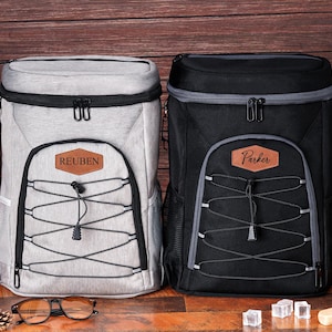May include: Two insulated cooler backpacks, one light gray and one black, with personalized leatherette name patches. Both have black zippers, gray trim, and a bungee cord design on the front. The light gray backpack has the name "Reuben" and the black one has the name "Parker".