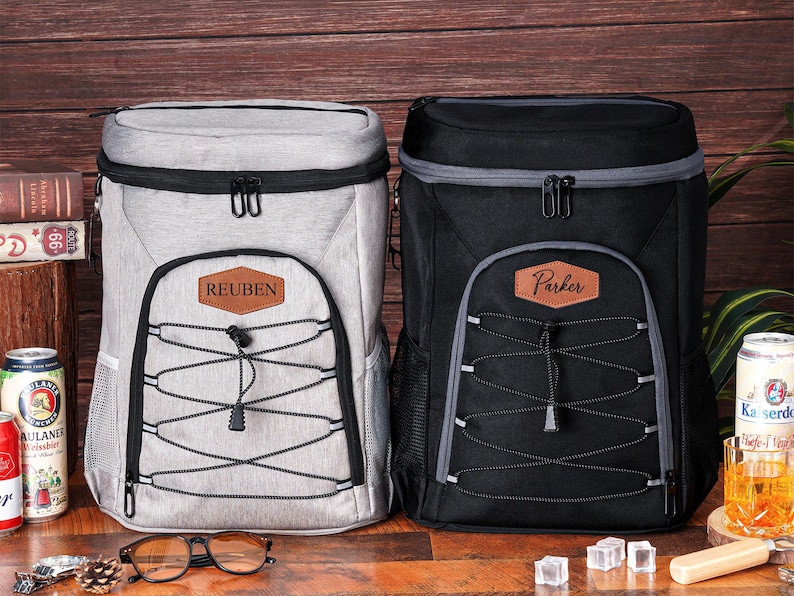 May include: Two insulated cooler backpacks, one light gray and one black, with personalized leatherette name patches. Both have black zippers, gray trim, and a bungee cord design on the front. The light gray backpack has the name "Reuben" and the black one has the name "Parker".