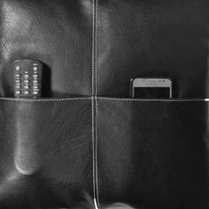May include: Black leather pillow with two pockets, a remote control in one pocket and a smartphone in the other. The pillow has white stitching details and a textured surface.