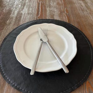 May include: A white plate with a scalloped edge, a knife, and a fork are arranged on a black placemat with white stitching. The placemat sits on a wooden table, creating a simple, elegant table setting.