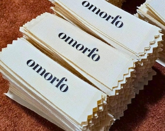 Könnte beinhalten: Ein Stapel rechteckiger, cremefarbener Stoffetiketten mit gezackten Rändern. Jedes Etikett trägt das Wort omorfo, das in einer fetten schwarzen Serifenschrift mittig aufgedruckt ist. Die Etiketten sind ordentlich übereinander gestapelt.