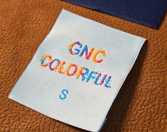 Puede incluir: Una etiqueta de tela blanca con el texto bordado "GNC COLORFUL" en un arcoíris de colores. Debajo del texto está la letra "S" en azul. Una etiqueta azul marino es visible en el fondo.