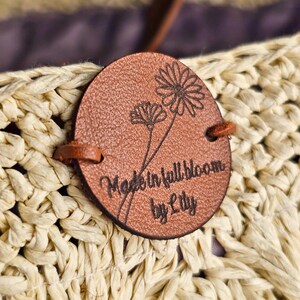 Può includere: Un'etichetta rotonda in pelle marrone con la scritta "Made in full bloom by Lily" e illustrazioni floreali. L'etichetta è attaccata a una borsa di paglia intrecciata, con un cinturino in pelle visibile.