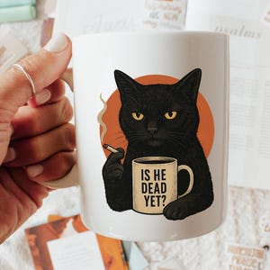 May include: White ceramic mug featuring a black cat illustration with yellow eyes, holding a cigarette and a mug that reads "IS HE DEAD YET?". The cat is set against an orange circle. A humorous and unique gift idea.