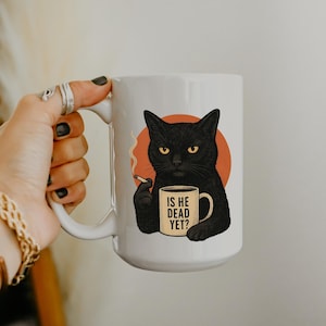 May include: White ceramic mug with a black cat illustration, yellow eyes, holding a cigarette and a mug that says "IS HE DEAD YET?". The cat is set against an orange circle.