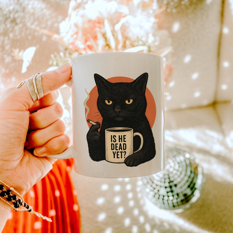 May include: White ceramic mug with a black cat illustration. The cat, with yellow eyes, holds a cigarette and a mug with the text "IS HE DEAD YET?". The cat is set against a peach-coloured circle. A humorous design.