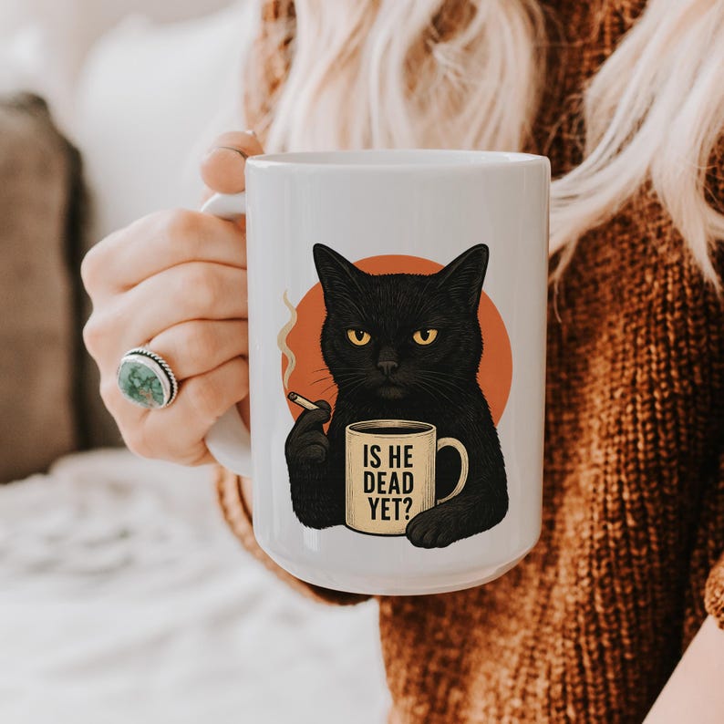 May include: White ceramic mug with a black cat illustration, yellow eyes, holding a cigarette and a mug that says "IS HE DEAD YET?". The cat is set against an orange circle.