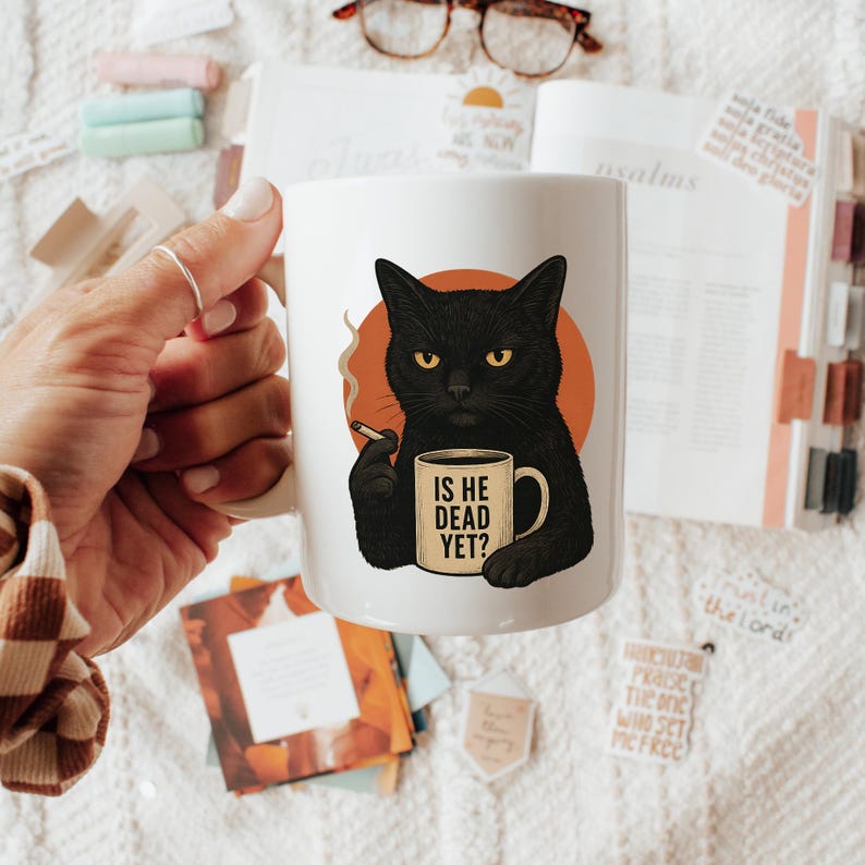 May include: White ceramic mug with a black cat illustration, holding a cigarette and a mug that reads "IS HE DEAD YET?". The cat is set against an orange circle. A humorous gift idea. The mug is a standard size.