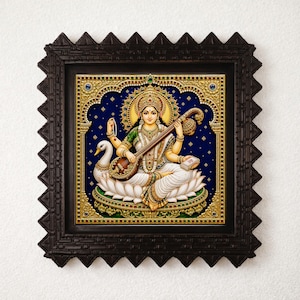 May include: A framed wall art piece featuring a detailed illustration of the Hindu goddess Saraswati seated on a white lotus flower. She holds a veena, a stringed instrument, and prayer beads, with a white swan positioned beside her on a dark blue background.