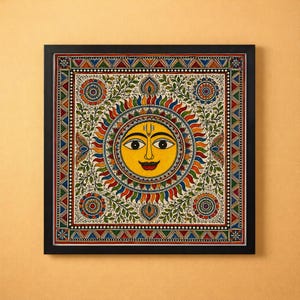 May include: A square framed wall art piece featuring a central yellow sun face with a red tilak, surrounded by colorful rays, intricate floral patterns, and geometric borders in red, blue, green, and orange hues on a cream-colored background.