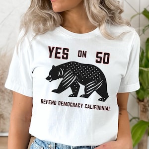 May include: White t-shirt with the text "YES ON 50" above a black bear graphic. Below the bear, the text reads "DEFEND DEMOCRACY CALIFORNIA!". The shirt is worn with blue denim jeans.