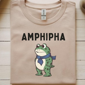 May include: A tan t-shirt with the word "AMPHIPHA" printed in black. Below the text is a cartoon frog wearing a blue scarf. The frog is green with dark green spots.