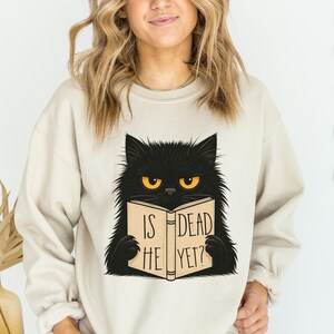 Pode incluir: Camisola bege com um gato preto de olhos amarelos lendo um livro. O livro diz "IS HE DEAD YET?". O gato tem pelo fofo e est&aacute; centralizado na camisola. A camisola tem mangas compridas e gola redonda.