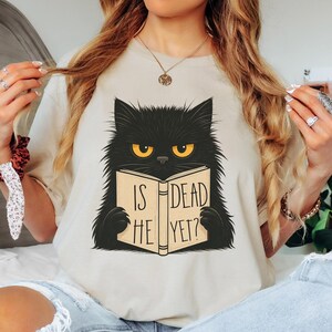 May include: Beige t-shirt featuring a black cat reading a book with the text "IS HE DEAD YET?" The cat has yellow eyes and a stern expression. The shirt is a casual style.