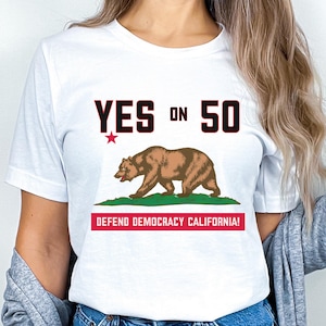 May include: White t-shirt featuring the California state flag with a brown bear, green grass, and the text "YES ON 50". Below the flag is the text "DEFEND DEMOCRACY CALIFORNIA!" in red.
